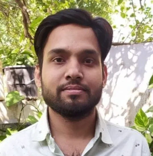 Brijaditya Sharma smiling outdoors in a light shirt, with green plants behind him
